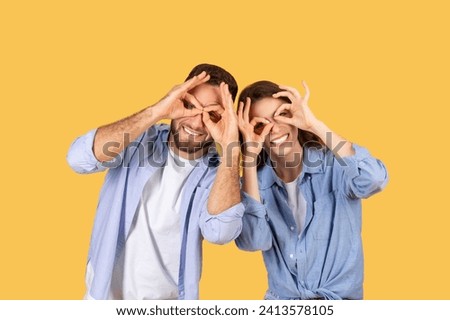 Laughing man and woman playfully making binocular shapes with their hands around their eyes, sharing fun moment on bright yellow backdrop
