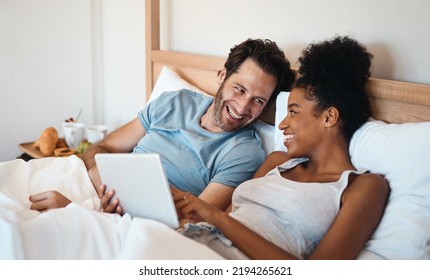 Laughing Married Couple With A Tablet In Bed Watching Funny Comedy Film Or Movie Together On The Weekend Indoors At Home. Loving, Smiling And Romantic Husband And Wife Looking At Meme On Social