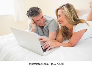 Laughing Mature Couple Lying On A Bed In A Bedroom Using A Laptop