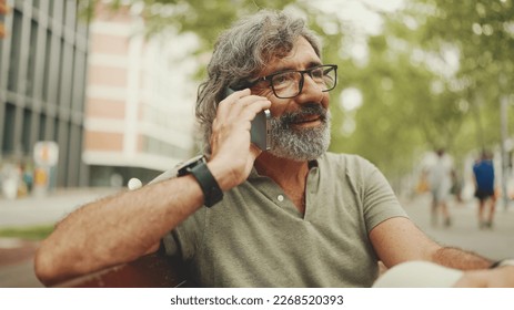 Laughing middle-aged man with gray hair and beard wearing casual clothes sits on bench and uses mobile phone. Mature gentleman in eyeglasses talking on smartphone - Powered by Shutterstock