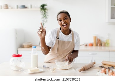 Laughing Millennial African American Woman In Apron Prepares Dough For Baking And Raises Finger Up In Light Scandinavian Kitchen Interior. Got Idea, Cooking Pizza, Pie At Home And Facial Expression