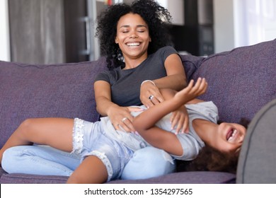 Laughing mixed race mother elder sister or young nanny tickling sweet little girl daughter family having fun at home sitting on couch playing together, weekend activities free time and leisure concept