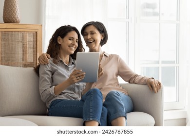 Laughing mother and 35s daughter enjoy new application, sit together on couch in living room using modern digital tablet device, watch video, multigenerational family having fun spend time on internet - Powered by Shutterstock