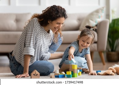 Laughing mother and little daughter wearing princess diadem playing colorful wooden blocks, constructing tower, sitting on warm floor with underfloor heating, family enjoying leisure time together - Powered by Shutterstock