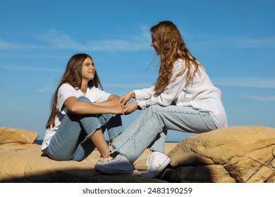 Laughing mother and teenage daughter, both with long hair, sit hand in hand on a rocky seashore against a clear blue sky. - Powered by Shutterstock