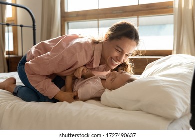Laughing mother tickling adorable little daughter lying on comfortable bed at home, family having fun in bedroom in the morning, happy mum and cute preschool girls playing funny game together