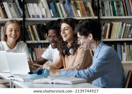 Laughing multi ethnic students use laptop studying or having fun using laptop sit at shared table in university library, having friendly relations, prepare for exams together using modern tech concept