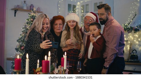 Laughing Multi Generational Family Gathering At Table And Waving Hands At Smartphone While Taking Selfie During Xmas Dinner At Home