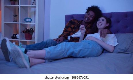 Laughing Multiethnic Family Watching Movie Together At Home In Evening. Diverse Young Couple Relaxing On Bed And Watching Comedy On Tv At Night In Bedroom