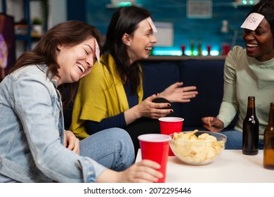 Laughing multi-ethnic friends playing who i am game using stickey notes enjoying spending time together during wekeend party at home. Group of people drinking beer guessing correct answer
