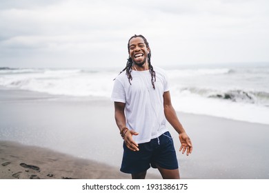 Laughing muscular African American male in white shirt and blue shorts with long dreadlocks and earphones standing on sandy beach with waving sea behind - Powered by Shutterstock