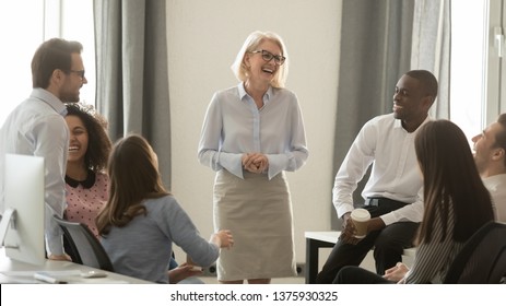 Laughing old coach team leader talking with diverse coworkers chatting at business meeting, friendly multi racial office workers and middle aged woman ceo have fun conversation at coffee break concept