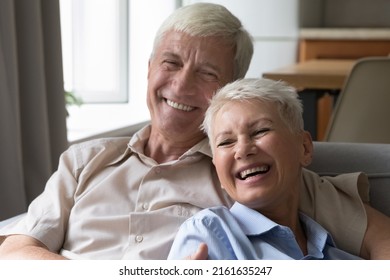Laughing older age married couple embrace on cozy sofa at living room having fun at home spend happy time on retirement. Smiling elderly spouses having good healthy teeth enjoy sweet moment together