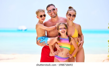 Laughing Parents With Boy And Girl In Vivid Swimsuits   Smiling At Camera Standing On Tropical Shoreline