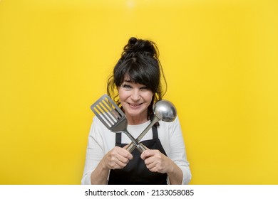 Laughing Senior Woman In Black Apron On Yellow Background Holding Crossed Scoop And Shovel, Seniors Activity Concept, Tasty Food And Cooking.