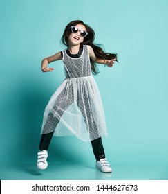 Laughing stylish asian baby girl kid in modern clothes and sunglasses jumps dances cool in gangnam style at party having fun makes show on blue mint background