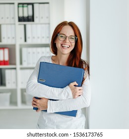 Laughing successful young businesswoman standing in a large airy office clutching a file in her arms grinning at the camera