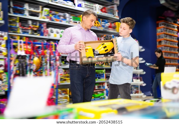 Laughing teenage boy with his father\
buying toy cement mixer machine in modern toy\
store