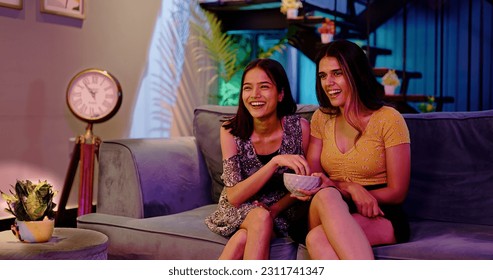 Laughing two Indian Girlfriend Sitting on sofa enjoying late night watching funny TV series at home. Female couple looking comedy movie on television eating popcorn snacks spend weekend time together - Powered by Shutterstock