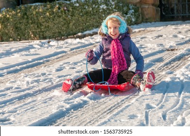 Laughing, Vibrantly Dressed Young Girl With Blue Ear Muffs Sledging Down A Slope