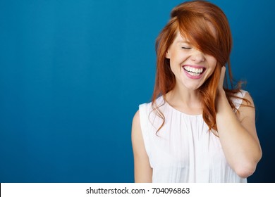 Laughing vivacious young redhead woman with a beaming smile holding her long red hair over one hand on blue with copy space