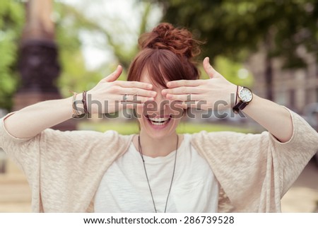 Laughing vivacious young woman holding her hands to her eyes as she stands outdoors in an urban park
