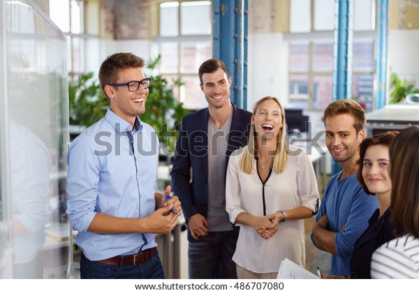 Laughing white collar workers standing\
around white board during meeting in small\
office