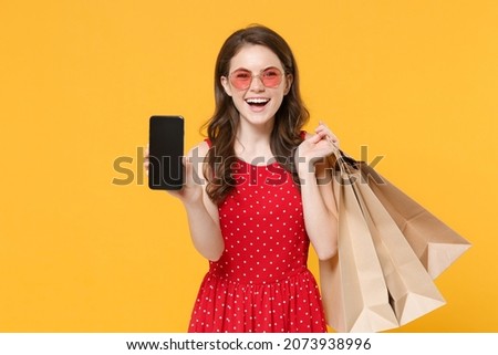 Laughing woman girl in red summer dress, eyeglasses isolated on yellow background. People lifestyle concept. Hold package bag with purchases after shopping, hold mobile phone with blank empty screen