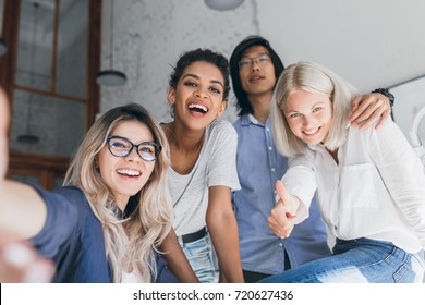 Laughing Woman In Jeans Posing Between European Lady And Asian Male Friend. Indoor Portrait Of International Students Having Fun In Campus And Making Selfie After Exams.