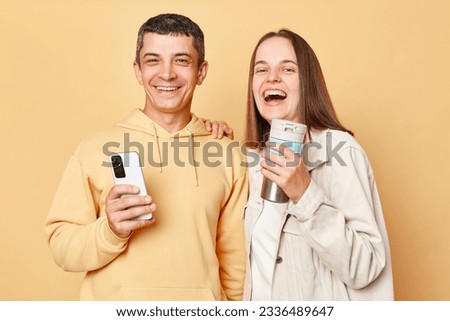 Laughing woman and man wearing casual style clothing standing isolated over beige background spending leisure time together using phone drinking hotbeverage.