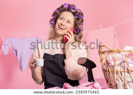 A laughing woman orders home cleaning services over the phone. Fed up with doing household chores, she sips coffee from a cup while holding rollers in hair. Portrait of a girl on a pink background.