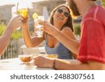 Laughing woman in sunglasses raises her drink in a toast during a lively outdoor gathering. Golden hour light bathes the scene, capturing the essence of carefree summer socializing with friends.