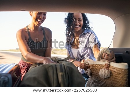 Laughing women unload the trunk of the car from the suitcases. Carefree multiethnic young women having fun during road trip in car on a sunset. Young latin woman enjoying road trip with her friend.