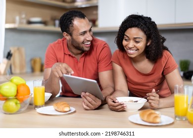 Laughing Young African American Couple Have Breakfast, Enjoy Chatting, Watching Video In Tablet And Good Morning In Kitchen Interior. New App And Online Call, Meeting Remote, Love And Relationships