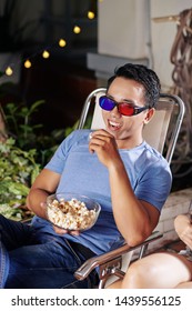 Laughing Young Asian Man In 3D Glasses Eating Popcorn And Watching Comedy Movie In Backyard