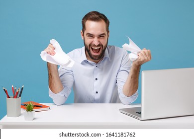 Laughing young bearded man in light shirt work at desk with pc laptop isolated on pastel blue background. Achievement business career concept. Mock up copy space. Screaming tearing paper documents