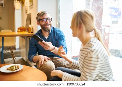 Laughing young bearded man showing his girlfriend funny video in smartphone while both spending time in cafe - Powered by Shutterstock