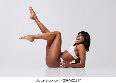 Laughing. Young beautiful dark skinned girl in white underwear lying on floor isolated over gray background. Wellness, wellbeing, fitness, fashion concept. Natural beauty of female body