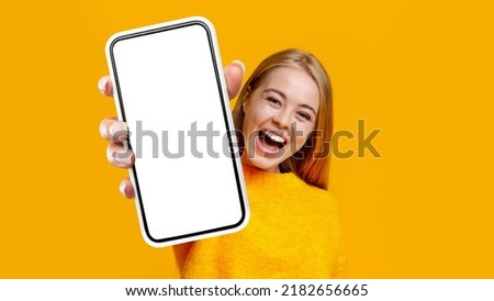 Laughing young blonde woman showing mobile phone with empty white screen, recommending newest entertaining mobile application, yellow studio background, mockup, copy space, collage