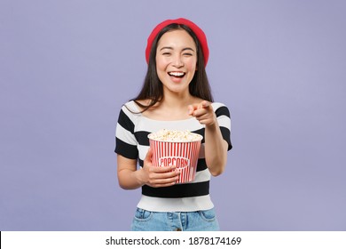 Laughing Young Brunette Asian Woman In Striped T-shirt Red Beret Watching Movie Film Hold Bucket Of Popcorn Pointing Index Finger On Camera Isolated On Pastel Violet Colour Background Studio Portrait