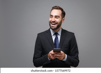 Laughing young business man in suit shirt tie posing isolated on grey background. Achievement career wealth business concept. Mock up copy space. Using mobile phone typing sms message, looking aside