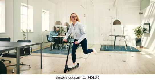 Laughing young businesswoman riding a scooter around a large modern office during her work break