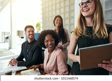 Laughing Young Businesswoman Standing In A Modern Office With A Diverse Group Of Work Colleagues Smiling In The Background