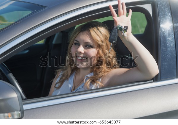 Laughing young driver\
woman is sitting in the car. The girl is waving at camera with car\
keys in her hand.