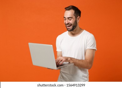 Laughing Young Man In Casual White T-shirt Posing Isolated On Bright Orange Background Studio Portrait. People Sincere Emotions Lifestyle Concept. Mock Up Copy Space. Working On Laptop Pc Computer