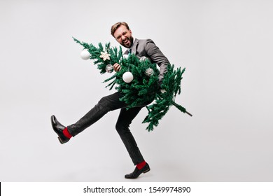 Laughing Young Man In Trendy Costume Enjoying New Year Party. Smiling Office Worker Holding Christmas Tree.