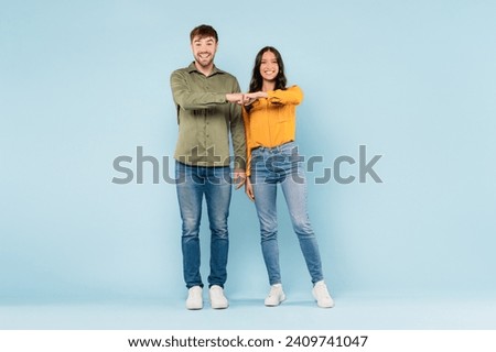 Laughing young man and woman in casual clothes fist bumping in friendly greeting or agreement, standing against soft blue background.