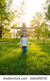 Laughing Young Mom And Her Small Daughter, With Open Arms, Are Walking Along The Green Meadow, Among Green Trees On A Sunny Evening. Happy Childhood In Nature. Back Light.