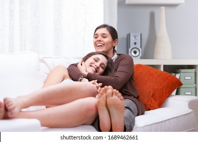 Laughing Young Mother And Daughter Relaxing Barefoot On A Comfortable Sofa In The Living Room Hugging And Smiling In A High Key Portrait With Copy Space