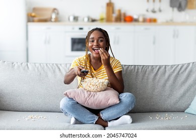 Laughing young pretty black woman with long braids sitting on sofa, eating popcorn and watching TV at home, holding remote controller, enjoying nice movie at weekend, copy space - Powered by Shutterstock